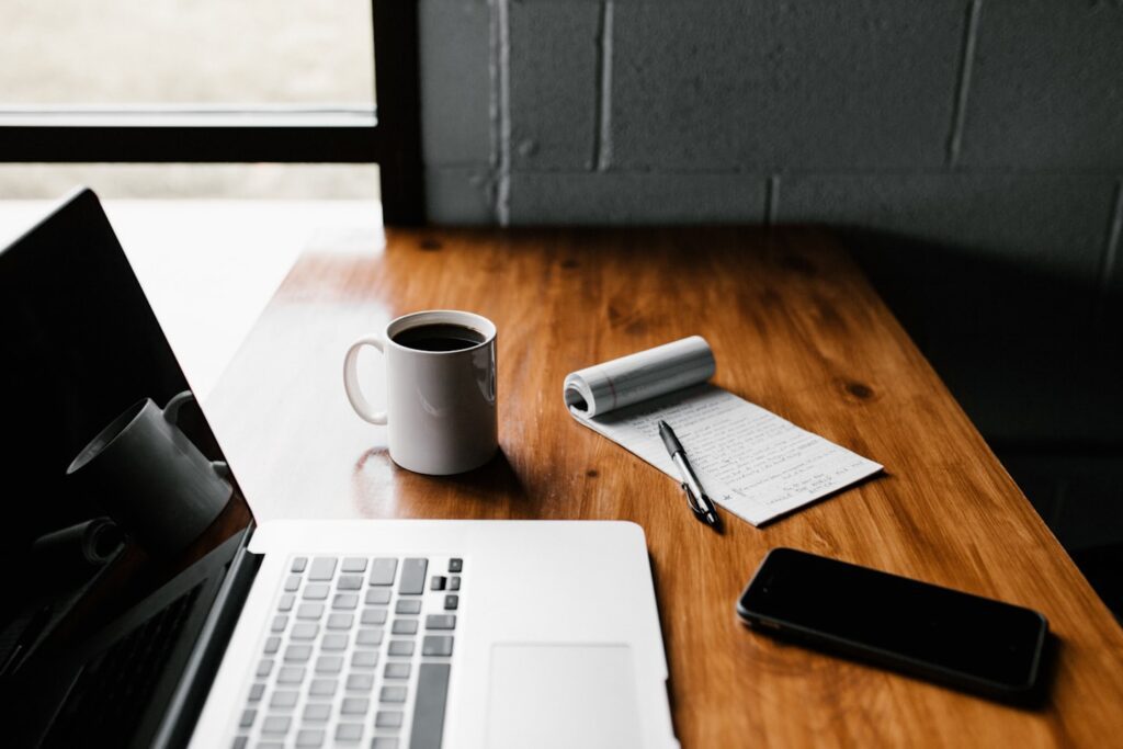 Vemos una laptop apagada sobre un escritorio de madera, al lado una taza de café y una libreta para tomar apuntes.